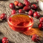 Recipiente de vidro com óleo de rosa mosqueta dentro e com frutos ao seu redor sobre uma mesa